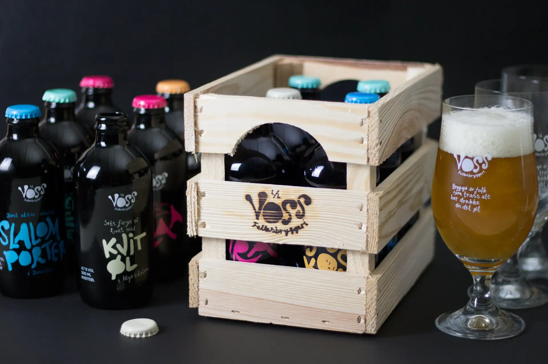 Voss Fellesbryggeri-flasker med fargede kapsler ved siden av en trekasse, med et fylt ølglass til høyre